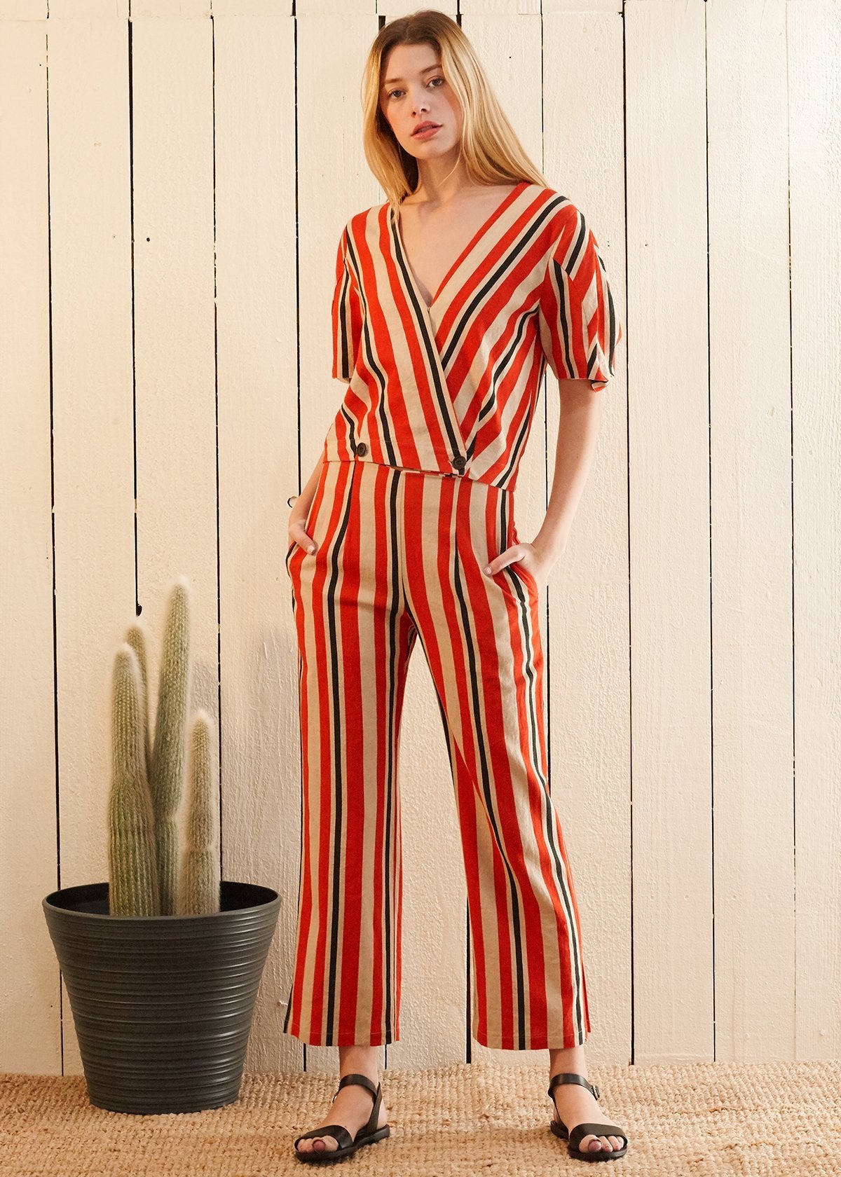 Woman wearing a red and white striped jumpsuit standing next to a potted cactus against a wooden wall.